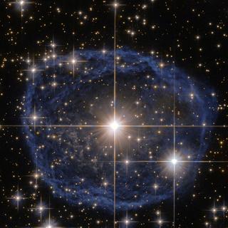 Uma grande bolha azul com uma estrela brilhante no centro contra um fundo preto repleto de estrelas, esta é uma bela imagem do Telescópio Espacial Hubble da NASA/ESA que mostra uma estrela Wolf-Rayet conhecida como WR 31a, localizada a cerca de 30.000 anos-luz da Terra, na constelação de Carina (A Quilha). A distinta bolha azul que parece circundar WR 31a é uma nebulosa Wolf-Rayet, uma nuvem interestelar de poeira, hidrogênio, hélio e outros gases. Criadas quando ventos estelares rápidos interagem com as camadas externas de hidrogênio ejetadas pelas estrelas Wolf-Rayet, essas nebulosas são frequentemente em forma de anel ou esféricas. A bolha, que se formou há cerca de 20 mil anos, está a expandir-se a uma velocidade de cerca de 220 mil quilómetros por hora! Infelizmente, o ciclo de vida de uma estrela Wolf-Rayet é de apenas algumas centenas de milhares de anos, um piscar de olhos em termos cósmicos. Embora as suas vidas tenham começado com uma massa pelo menos 20 vezes a do Sol, as estrelas Wolf-Rayet normalmente perdem metade da sua massa em menos de 100.000 anos. E o WR 31a não foge à regra. Terminará, portanto, a sua vida numa supernova espectacular, e a matéria estelar expelida pela sua explosão alimentará mais tarde uma nova geração de estrelas e planetas. © ESA/Hubble e NASA, Agradecimentos: Judy Schmidt