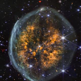 Uma faixa escura distinta entre duas nuvens cósmicas reforça a aparência cerebral da nebulosa PMR 1. O instrumento NIRCam (câmera infravermelha próxima) do Telescópio Espacial James Webb revela em uma única imagem várias fases das erupções de uma estrela no final de sua vida: a bolha externa esbranquiçada, em forma de crânio, provém de uma ejeção inicial, principalmente de hidrogênio, seguida por outros materiais mais pesados, visíveis em laranja no interior da nebulosa. Como acontece frequentemente nas imagens NIRCam, muitas estrelas, mesmo galáxias distantes, são visíveis atrás da nebulosa. Além da sua aparência singular, o PMR 1 ainda esconde muitos mistérios. Não está claro se a estrela atrás da nebulosa tem massa suficiente para explodir como uma supernova, ou se evoluirá para uma anã branca densa assim que as suas camadas exteriores forem ejetadas. © NASA, ESA, CSA, STScI, Processamento de imagem: Joseph DePasquale (STScI) 